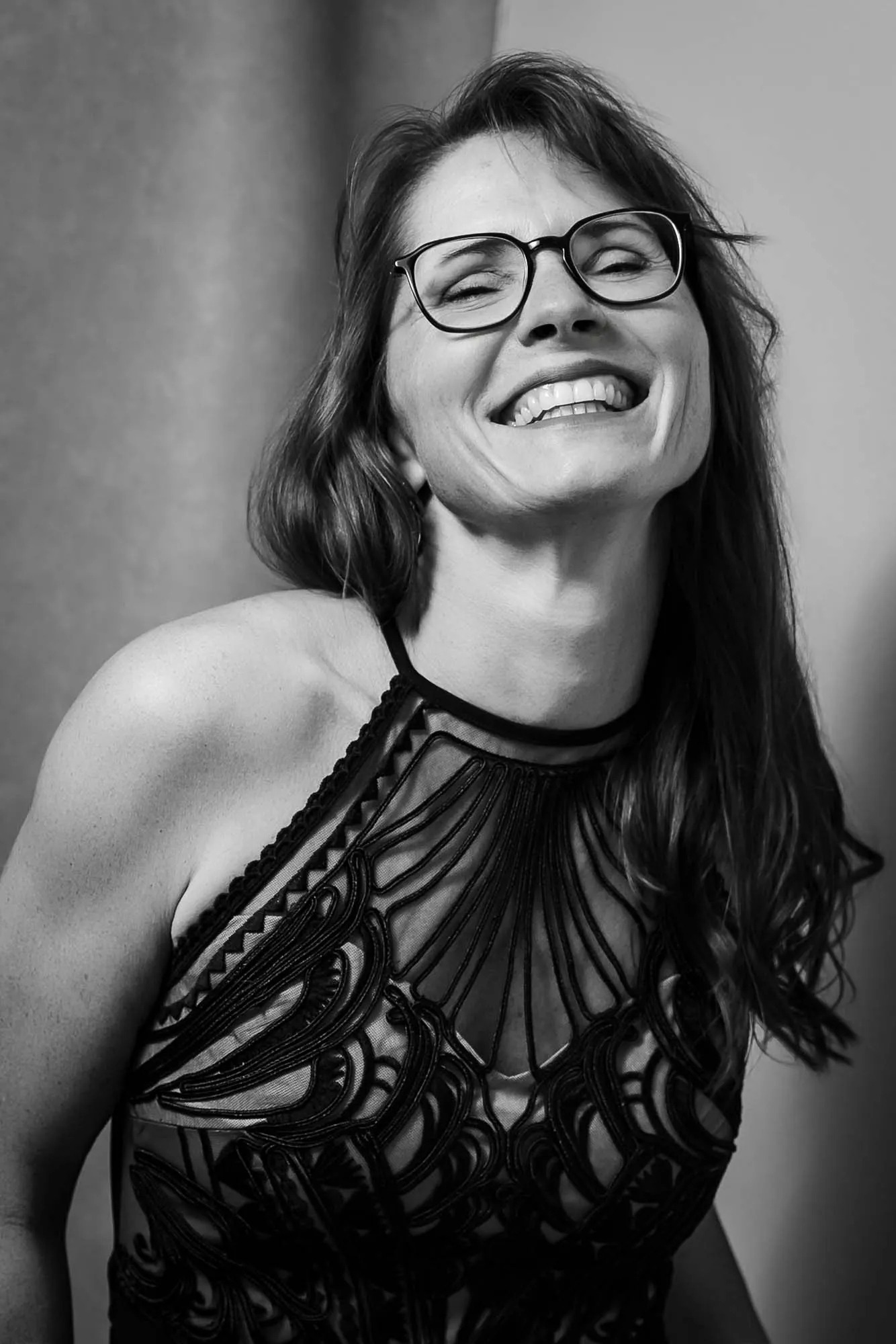 Frau mit Brille im schwarzen Spitzenkleid lacht mit geschlossenen Augen, Schwarz-Weiß-Porträt