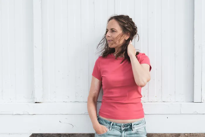 Juliane Klüß in rotem T-Shirt lehnt an einer weißen Holzwand, der Wind spielt in ihren Haaren
